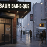 News crews photograph the darkened Dinosaur Bar-B-Que storefront in Brooklyn at dusk, with a “CLOSED” sign on the door and wet streets reflecting the sign. The scene captures the moment dinosaur bar-b-que closes restaurants, drawing media and sparking nationwide attention.