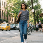 Katie Holmes walks a sunny New York sidewalk wearing a navy Breton-stripe top, straight-leg jeans, and white sneakers, carrying a brown tote past brownstones and yellow taxis with café tables in the background.
