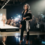 Miss J Alexander stands confidently on a fashion runway wearing a black velvet blazer with gold embroidery.