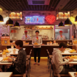 A colorful restaurant storefront decorated with large anime character decals and a vibrant banner announcing the Popeyes One Piece Collab to passing customers. Fans line up outside the entrance, which features a life-sized cutout of a pirate captain holding a fried chicken bucket.