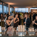 A crowded rooftop venue at dusk with product displays on a glass table in the foreground. Attendees mingle near floor-to-ceiling windows overlooking the city skyline.