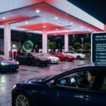 Night view of Tesla virtual queue supercharger system with digital queue board displaying wait times. Multiple Tesla vehicles wait in numbered stalls under illuminated canopy at busy station.